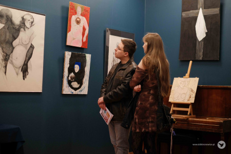 Mężczyzna w brązowej kurtce i kobieta w brązowej bluzce oglądają wystawę. Przyglądają się obrazowi, który przedstawia mężczyznę i kobietę.