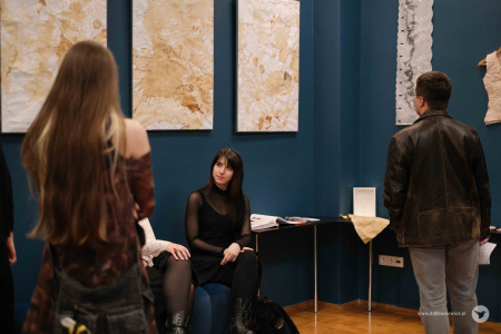 Dwie młode kobiety siedzą przy granatowej ścianie. Nad nimi zawieszone biało-beżowe obrazy. Obrazy są abstrakcjami.