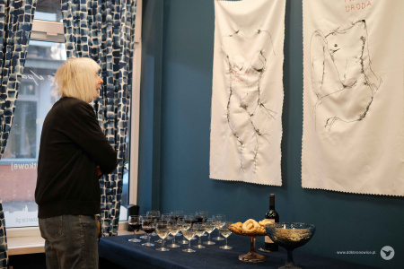 Kobieta ogląda dwa wyszywane obrazy przedstawiające sylwetki kobiet. Między nią, a ścianą z obrazami stoi stół. Na stole kieliszki, butelka wina i przekąski.