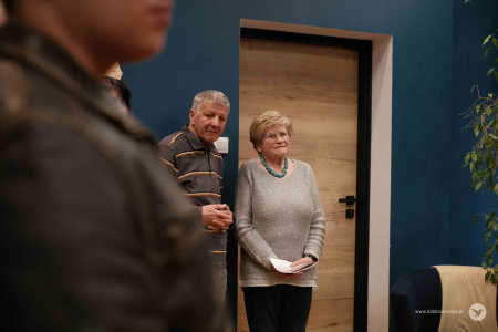 Starszy mężczyzna w bluzce w paski stoi obok starszej kobiety. Kobieta ubrana jest w szary sweter.