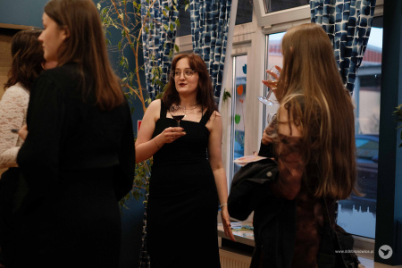 Na pierwszym planie dwie kobiety. Nie widać twarzy tych osób. W środku stoi młoda kobieta w czarnej sukience. W ręce trzyma kieliszek z napojem.