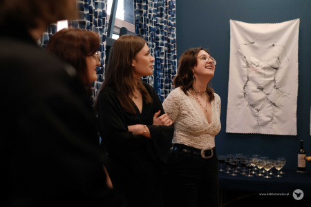 Trzy kobiety stoją obok siebie. Dwie z nich ubrane są na czarno. Kobieta stoją po prawej stronie w białej bluzce uśmiecha się. W tle widać białe wyszywane obrazy z wizerunkiem kobiet.