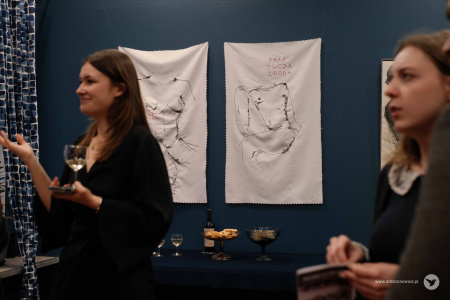 Na pierwszym planie dwie kobiety. Kobieta po lewej stronie trzyma kieliszek z napojem. Kobieta po prawej stronie trzyma broszurę dotyczącą wystawy. W tle widać dwa wyszywane obrazy z wizerunkiem kobiet.