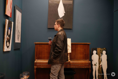 Mężczyzna w brązowej kurtce ogląda wystawę. W ręce trzyma kieliszek z napojem. W tle widać obraz przedstawiający krzyż i pianino.