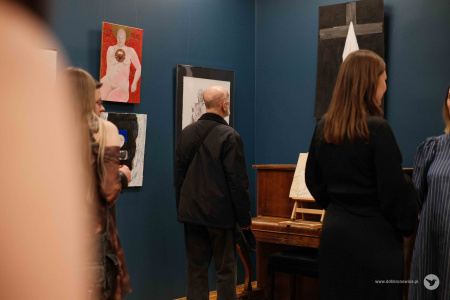 Na pierwszym planie ludzie oglądający wystawę. W środku mężczyzna ogląda obraz przedstawiający krzyż. Za nim widać czerwony obraz.