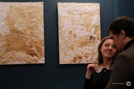 Po prawej stronie kobieta rozmawia z mężczyzną w brązowej kurtce. W tle wiszą biało-beżowe abstrakcje.