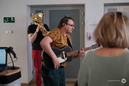 Mężczyzna w żółtej koszulce gra na gitarze. Za nim mężczyzna gra na euphonium. W tle wejście na sale teatralną.