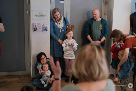 Na pierwszym planie kobieta klaszcze w dłonie. W tle mężczyzna w zielonej koszulce i niebieskiej bluzie stoi oparty o ścianę. Obok niego stoi kobieta w niebieskim płaszczu. Przed kobitą stoi dziewczynka w jasnej koszulce i brązowymi uszami na głowie.