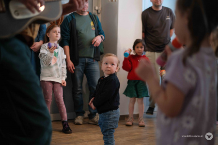 Dzieci trzymają w rękach własnoręcznie zrobione kolorowe grzechotki. W środku stoi mały chłopiec w czarnej bluzie i dżinsach.