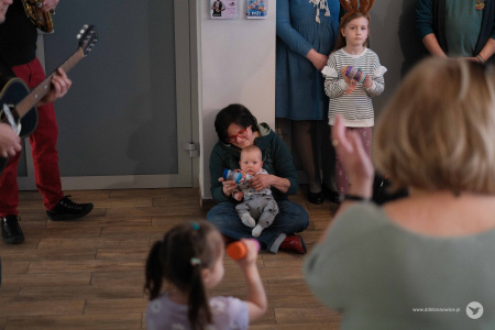 Po lewej stronie stoją muzycy. Po prawej stronie widać dziewczynkę. Na głowie ma opaskę z brązowymi uszami. Pośrodku przy ścianie siedzi kobieta i trzyma na nogach małe dziecko.