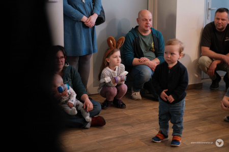 Na środku stoi mały chłopiec ubrany w czarną bluzę. Po lewej stronie kobieta trzymająca na nogach małe dziecko. W tle widać mężczyznę w zielonej koszulce.