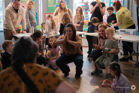 Sylwia Lasok prowadzi zajęcia dla dzieci. Muzyczka kuca i składa ręce. Wokół niej siedzą dzieci. W tle dorośli stoją przy białym stole warsztatowym.