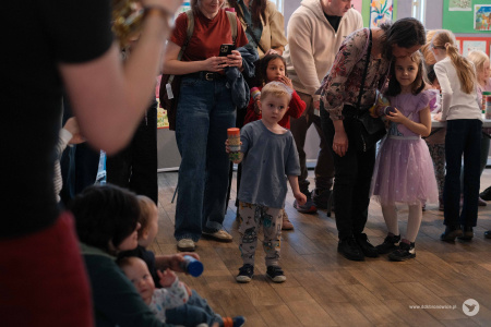Chłopiec w szarej koszulce pokazuje zrobioną kolorową zabawkę. Obok niego stoi dziewczynka w fioletowej sukience. W tle widać stojące osoby dorosłe.