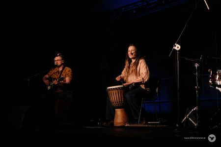 Sylwia Lasok siedzi na środku sceny na krześle. Obok niej gitarzysta w żółtej koszulce gra na gitarze. Wokalistka trzyma bęben między nogami.