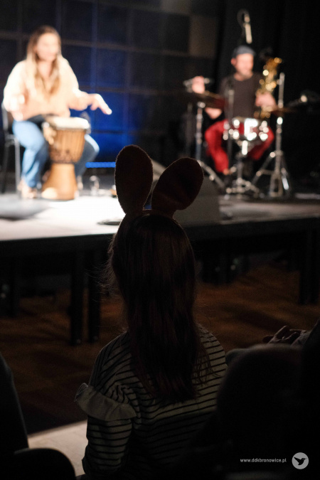 Na pierwszym planie widać dziewczynkę. Na głowie ma opaskę ze stojącymi brązowymi uszami. W tle muzycy graja koncert.