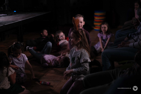Między sceną a pierwszym rzędem dzieci leża na podłodze. Śmieją się i uśmiechają.