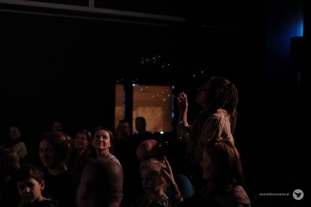 Sylwia Lasok puszcza bańki mydlane na dzieci i widownie. Kobieta na widowni wyraźnie się uśmiecha.