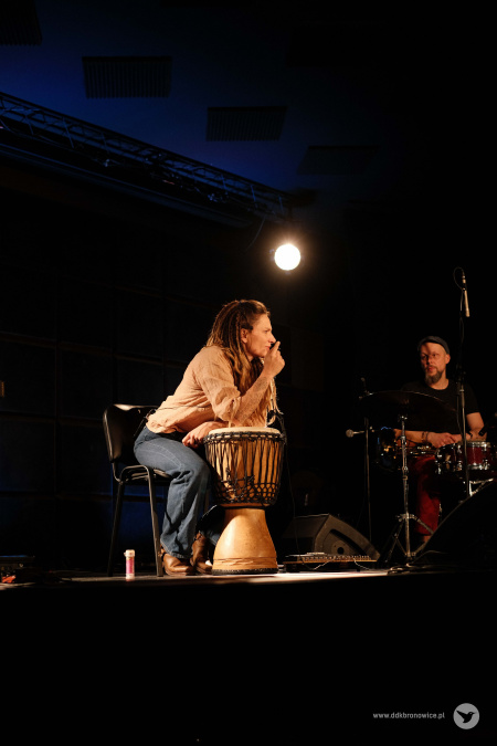 Scena. Na scenie Sylwia Lasok siedzi na krześle. Ciałem opiera się na stojący przed nią bęben. Zwraca się do publiczności. Po prawej stronie widać perkusistę w czarnej koszulce.