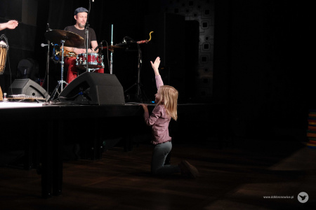 Pod sceną stoi dziewczynka. Ubrana jest w szare spodnie i różową koszulkę. Uśmiecha się i macha do muzyków.