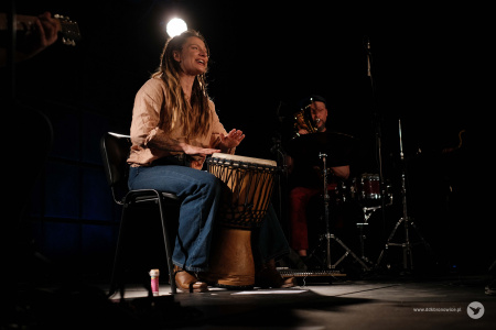 Na scenie Sylwia Lasok gra na bębnie. Śpiewa do mikroportu. Siedzi na czarnym krześle pośrodku sceny.