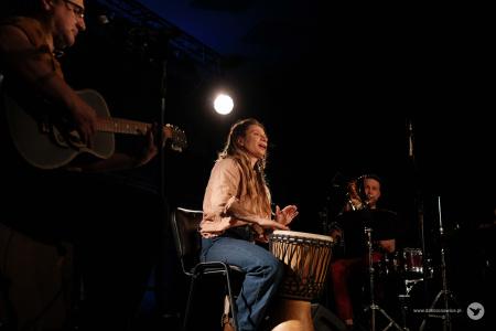 Na scenie Sylwia Lasok gra na bębnie. Śpiewa do mikroportu. Siedzi na czarnym krześle pośrodku sceny. Po prawej stronie mężczyzna gra na euphonium. Po lewej widać gitarzystę.