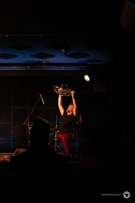 Muzyk w czarnej koszulce i czerwonych spodniach podnosi euphonium do góry.