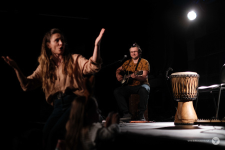Po prawej stronie na scenie oświetlony bęben. Obok niego siedzi uśmiechnięty gitarzysta. Pod sceną stoi Sylwia Lasok, która zwraca się w kierunku perkusisty.