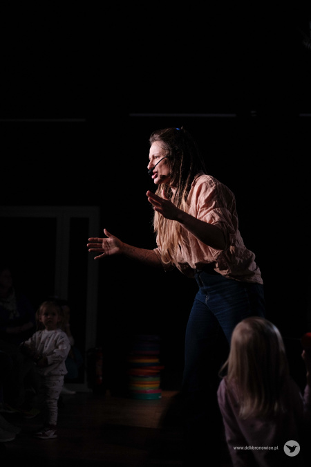 Wokalistka śpiewa piosenkę. Ma otwarte oczy i podniesione ręce. Wokół niej widać dzieci.