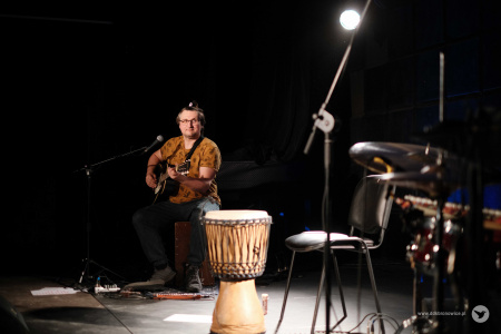 Na środku sceny podświetlony bęben. Za nim siedzi muzyk grający na gitarze. Patrzy się uśmiechnięty w kierunku perkusisty.