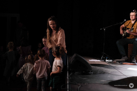 Artystka stoi pod sceną. Ma zamknięte oczy. Dzieci wokół niej się bawią.