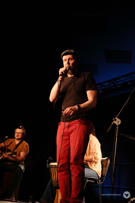 Na przodzie sceny stoi mężczyzna w czarnej koszulce i czerwonych spodniach. Trzyma w prawej ręce mikrofon. W tle widać Sylwię Lasok i gitarzystę w żółtej koszulce.