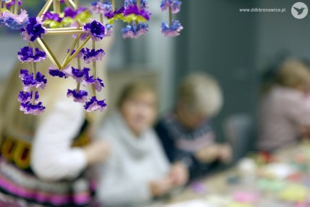 Z sufitu zwisa ludowy kolorowy pająk. Konstrukcja ma słomiane patyczki i kolorowe kwiatki. W tle widać pracujących uczestników warsztatów. Tło jest rozmazane.