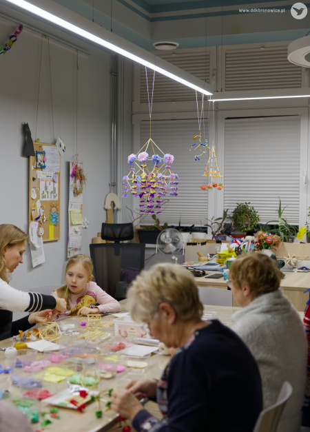 Przy stole warsztatowym pracują uczestnicy. Po lewej stronie widać dziewczynkę w różowej bluzce i kobietę, która jej pomaga. Po prawej stronie dwie starsze uczestniczki warsztatów. Z sufitu zwisają ludowe pająki.