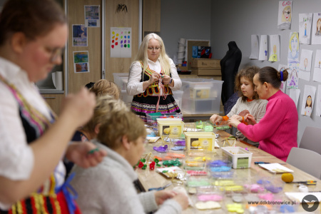 Pracownie Kultury Dziesiąta. Na stole warsztatowym kolorowa bibuła, nożyczki, koszyczki i artykuły papiernicze. Za stołem siedzi prowadząca warsztaty Iwona Barecka. Ubrana jest w strój ludowy. Przy stole siedzą uczestnicy warsztatów.