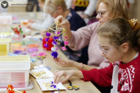 W tle kobieta tworzy niebiesko-fioletową ozdobę. Na pierwszym planie dziewczynka w czerwonym swetrze tworzy ludowego pająka z niebieskimi płatkami.