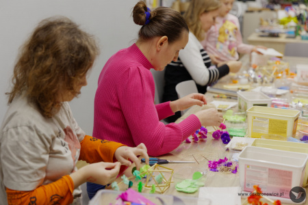 Przy stole, kobieta w różowym swetrze tworzy ludową ozdobę.