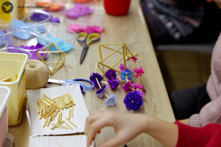 Mała konstrukcja leży na stole. Płatki są koloru różowego i niebieskiego. Na stole leżą też nożyczki, włóczka i materiały potrzebne do wykonania ozdoby.