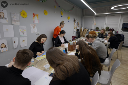 Uczestnicy malują obrazy przy stole warsztatowym.