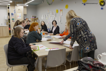 Po prawej stronie prowadząca warsztaty. Za stołem siedzą uczestnicy i malują obrazy.