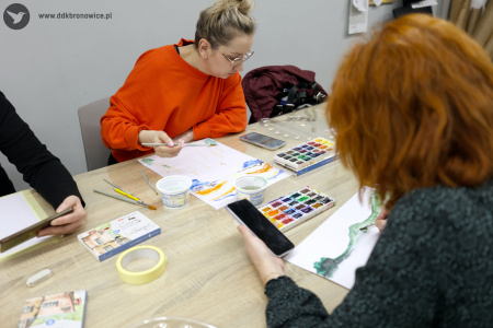 Za stołem siedzi uczestniczka w pomarańczowym swetrze. Na telefonie ma włączony obraz, który maluje. Obraz jest jest w kolorystyce pomarańczowo-niebieskiej.