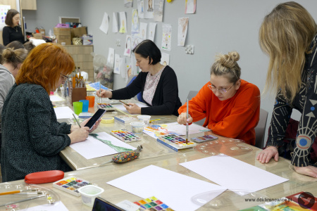Uczestniczka w pomarańczowym swetrze maluje obraz. Obok niej stoi prowadząca warsztaty. W tle uczestniczka w granatowym swetrze maluje swój obraz.