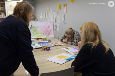 Uczestniczka w czarnej bluzie rozmawia z prowadzącą. Prowadząca ma przed sobą na stole niebiesko-żółty obraz.