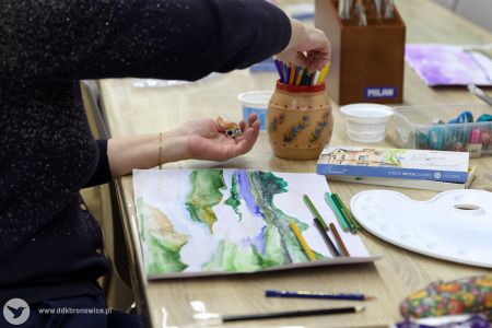 Uczestniczka maluje obraz. Obraz przedstawia zielone kontury wzgórz. Na stole oprócz akwareli leża kubeczki z kredkami.