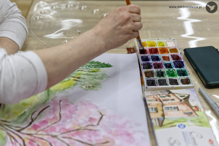 Uczestniczka w białej bluzce maluje obraz. Obraz przedstawia drzewa. Największe drzewo jest koloru różowego. W tle widać kilka mniejszych zielonych.