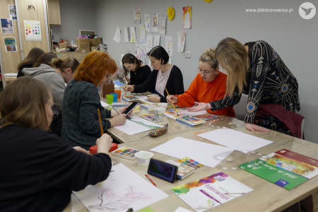 Uczestnicy malują obrazy akwarelą. Obok uczestniczki w pomarańczowym swetrze stoi prowadząca.
