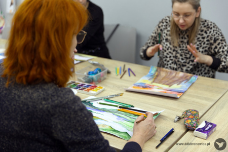 Naprzeciwko siebie siedzą dwie uczestniczki. Malują obrazy. Pierwszy obraz przedstawia zielone góry, a drugi jest w kolorystyce brązowo-niebieskiej.