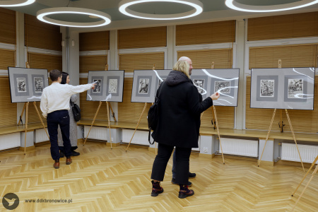 Obrazy wystawione na dużych sztalugach. Na jednej sztaludze wiszą dwa obrazy na szarych tłach. Obrazy ustawione są wzdłuż okien.