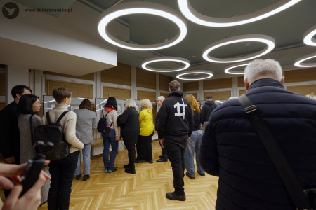 Duże grupa uczestników ogląda obrazy, ustawione wzdłuż dużych białych okien.