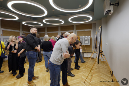 Mężczyzna w szarej bluzie i kobieta w czarnej bluzce przyglądają się dwóm obrazom na sztaludze. W tle inni uczestnicy oglądają obrazy.