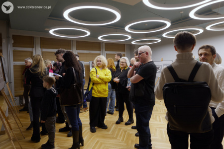 Uczestnicy wystawy oglądają obrazy. Mężczyzna w krótkiej koszulce przygląda się obrazom. Po lewej stronie rozmawiają dwaj mężczyźni w białych swetrach.